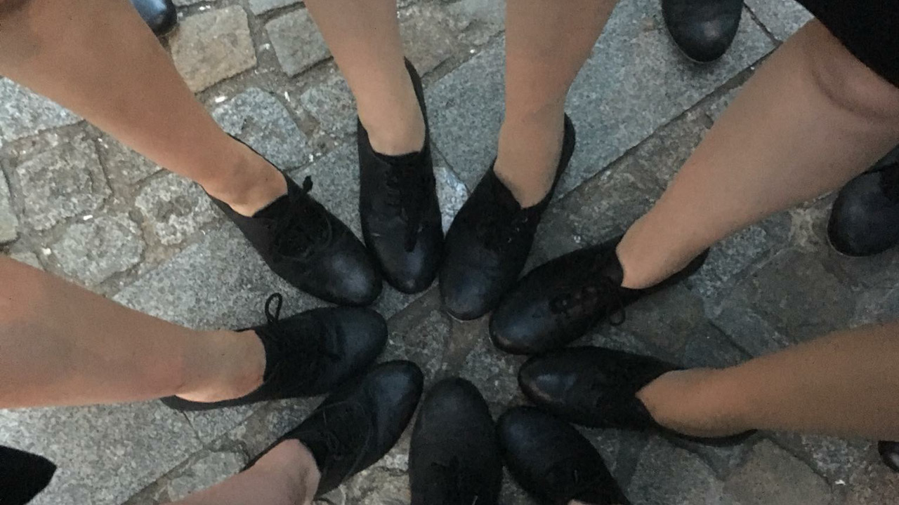 cirle of people each with one leg extended, all wearing black clogs  