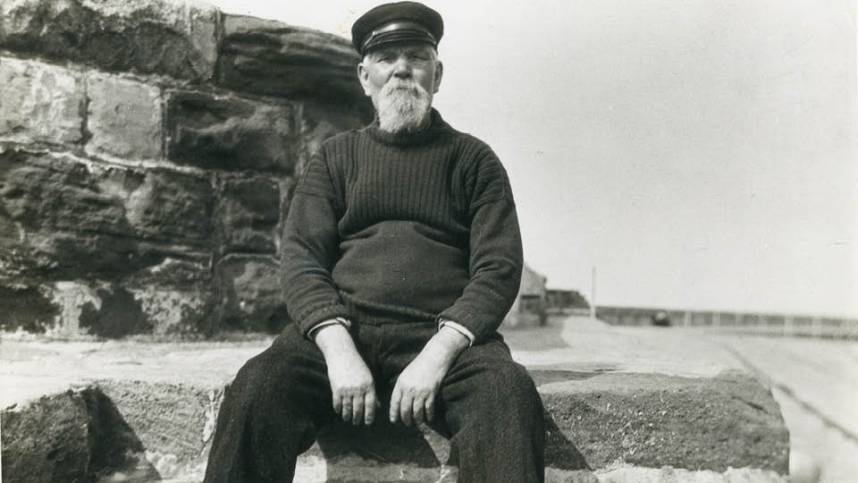 Black and white portrait of white-bearded sea dog wearing a cap and woolen jumper and sitting on a harbour wall